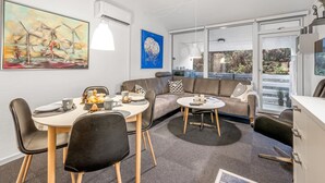 Una sala de estar moderna con comedor, un sofá y una mesa de centro. Hay un cuadro en la pared, una ventana con vistas y un ventilador de techo.