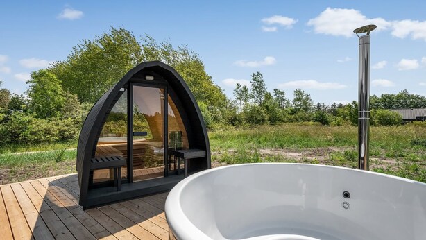 Un bain à remous installé sur une terrasse en bois, avec un espace salon clos par des baies vitrées et une structure en forme de pergola.