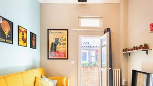 Un salon avec un canapé jaune, une affiche encadrée, un téléviseur et une porte donnant sur un balcon agrémenté de plantes.