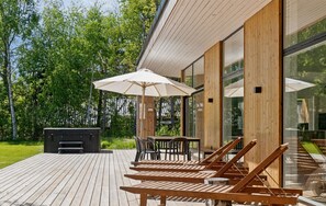 Una terraza con sillones de madera, sombrillas, una parrilla y un edificio con paneles de madera y paredes de vidrio.