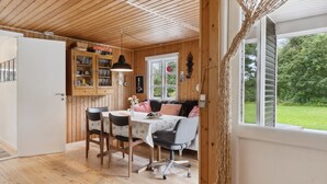 Een knus houten interieur met een eettafel en stoelen, een bank en een raam met uitzicht op een tuin.