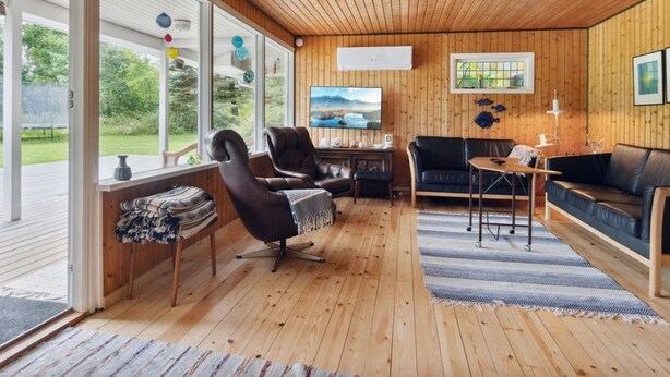 Een ruime woonkamer met houten vloeren en wanden, een groot raam met uitzicht op de omgeving, een televisie en een gezellige zithoek met een bank en fauteuils.