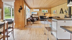 Een modern interieur met houten lambrisering, een eethoek met tafel en stoelen, een zithoek met een bank en televisie, en een keuken met een fornuis en kasten.