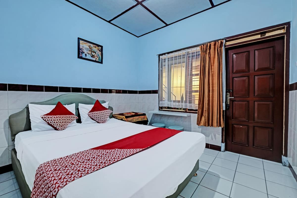 A hotel room with a bed, a window with curtains, a door, and a small table with a lamp.