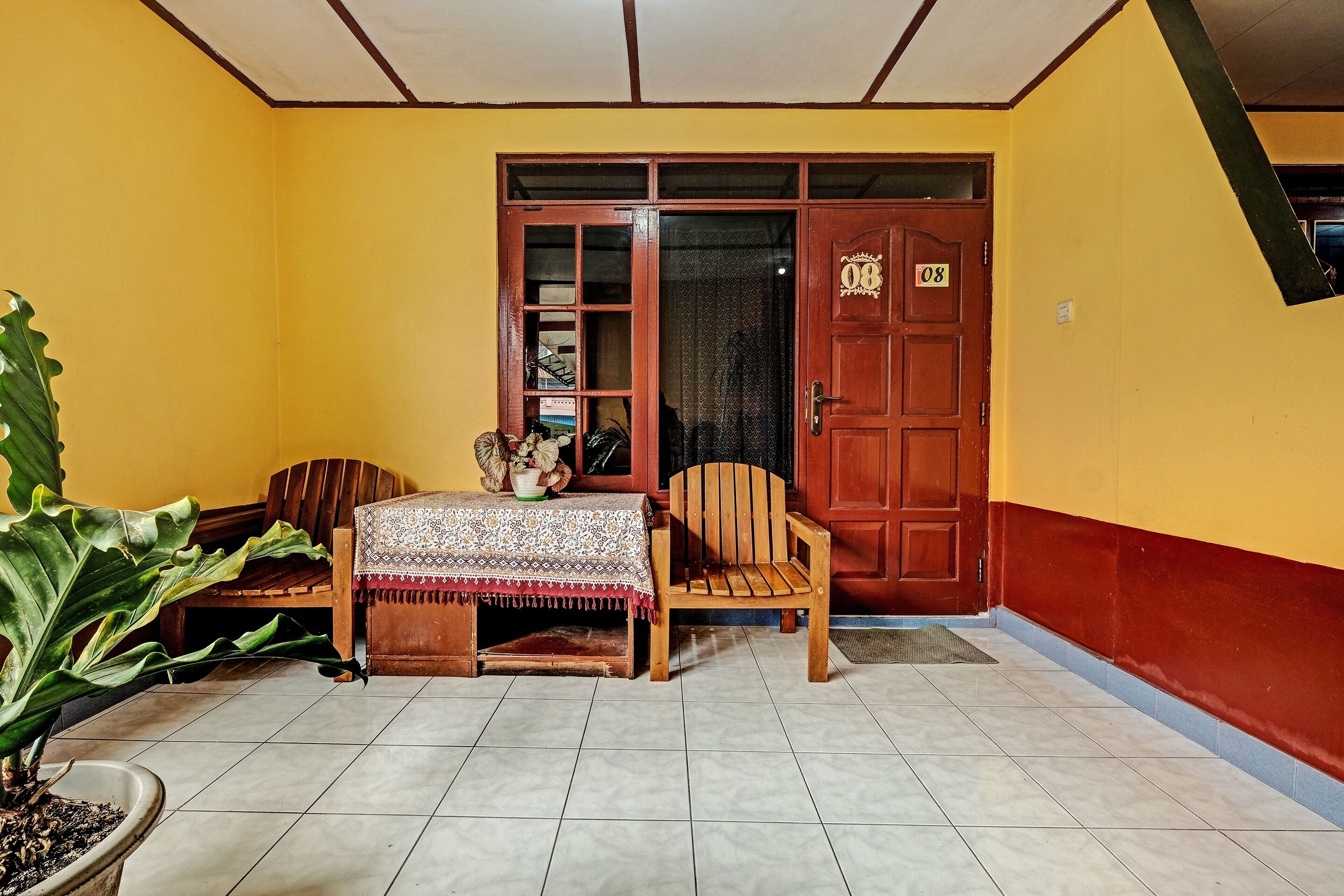 Una habitación con suelo de baldosas, una puerta de madera con placa de número, una silla de madera, una mesa con mantel estampado y una planta en maceta.