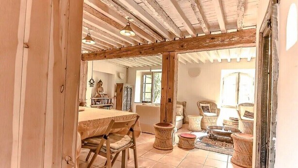 Un interior rústico con vigas de madera a la vista, una zona de comedor con mesa y sillas de madera, mobiliario de mimbre y vistas a un jardín por una ventana.