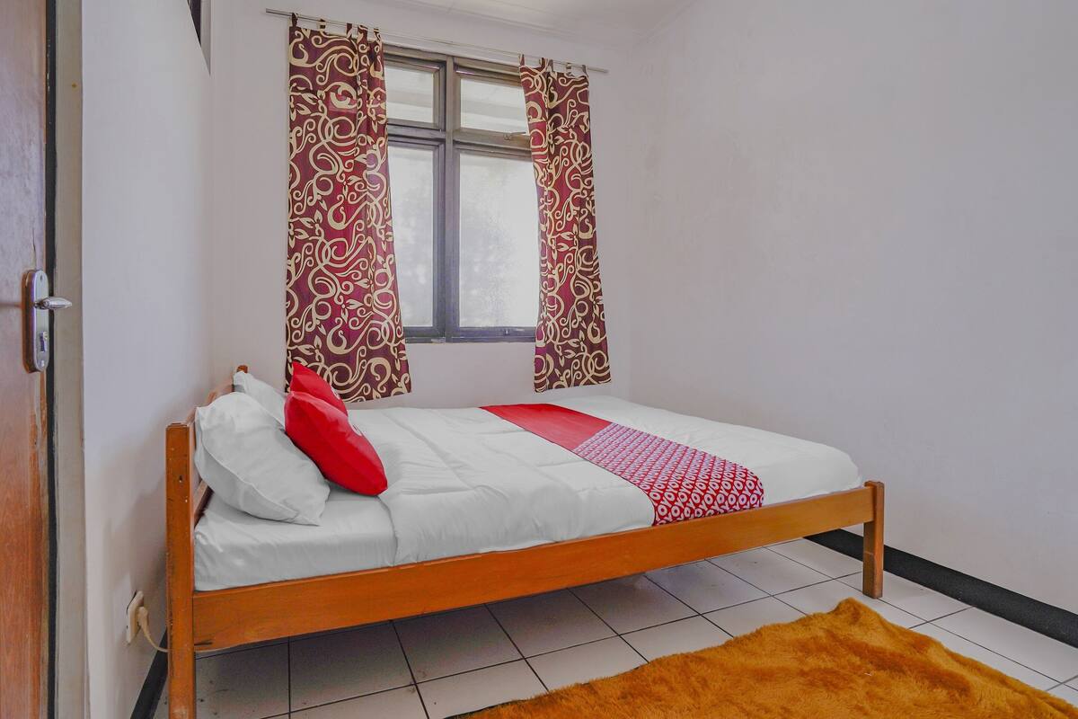 Uma cama de solteiro com estrutura de madeira, roupa de cama branca e almofada vermelha. Há uma cortina estampada na janela e um tapete castanho no chão.