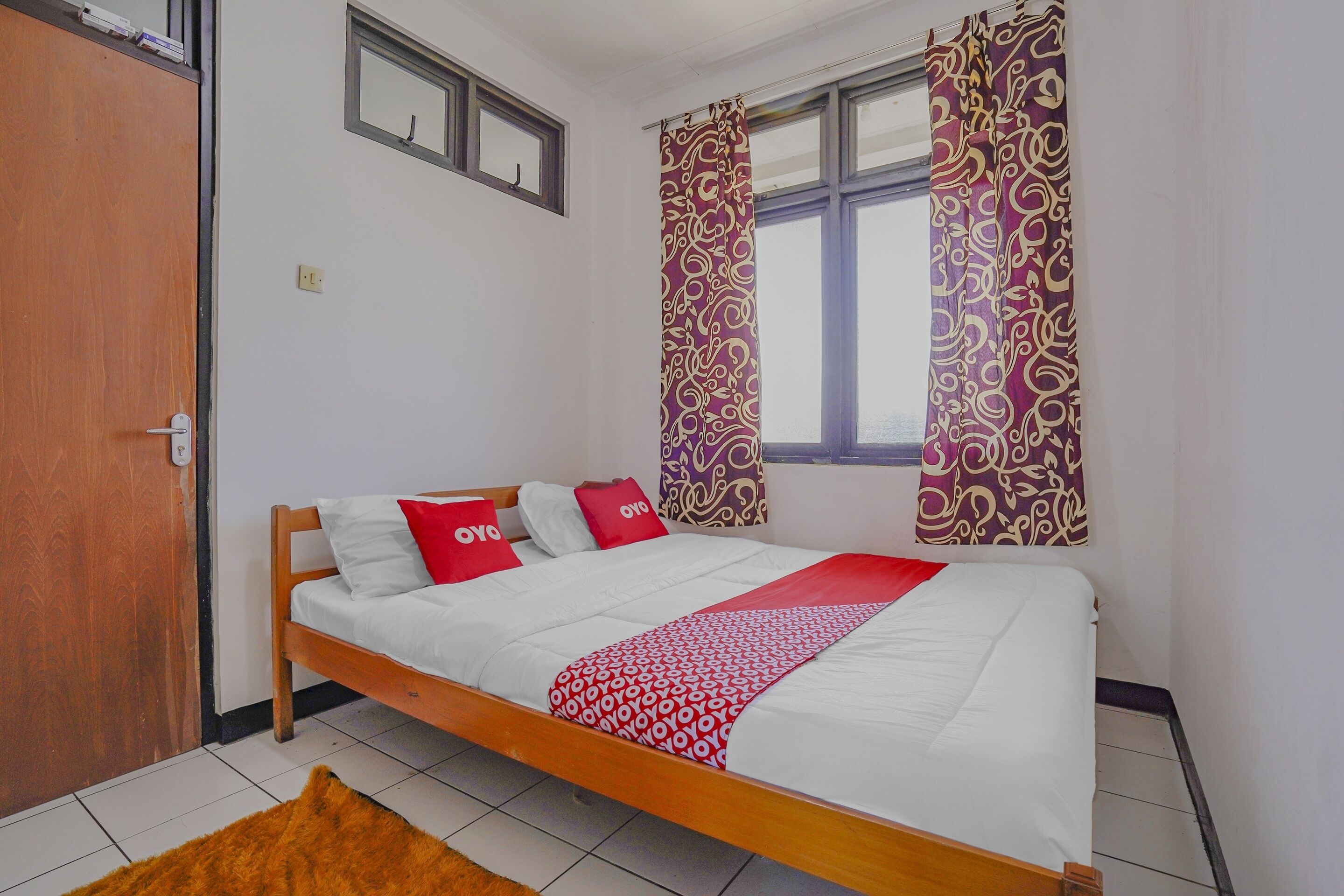 A single-bed room with a wooden frame, white bedding, red and white pillows, patterned curtains, and a window with blinds.