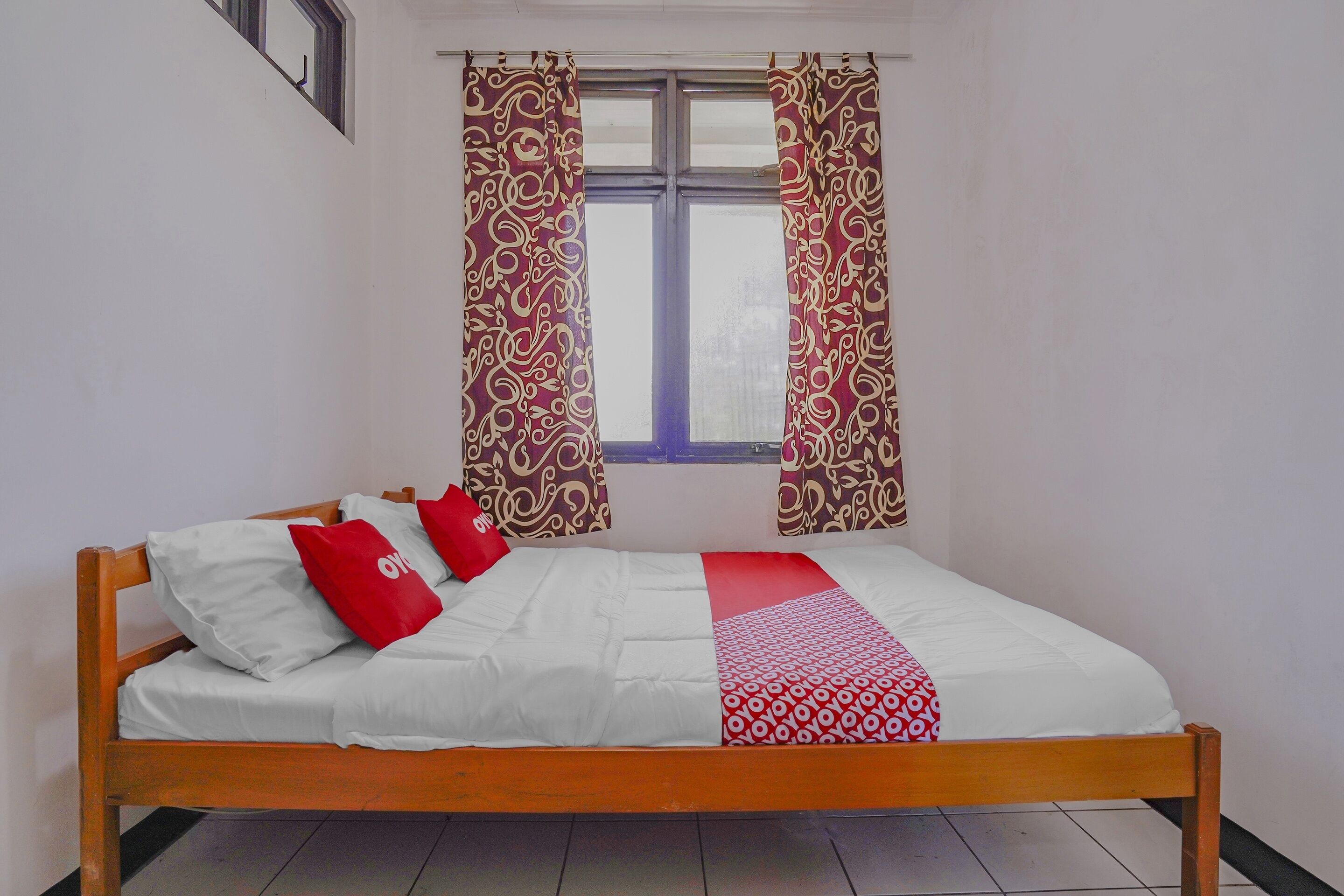 A single bed with a wooden frame, white bedding, and red pillows. There are patterned curtains on the window.