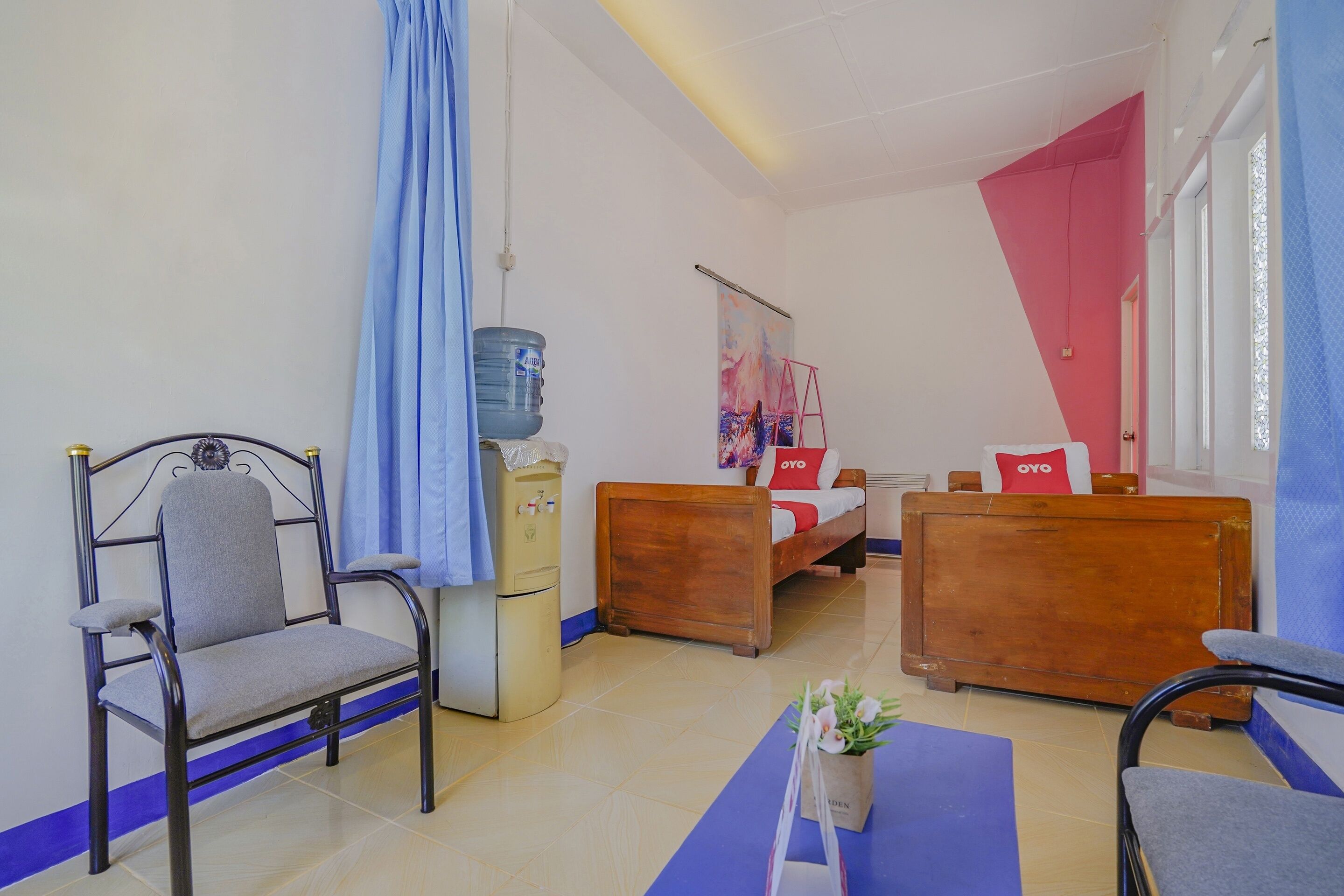 A room with a bed, two wooden nightstands, a desk, a chair, a water dispenser, and a wall-mounted shelf.