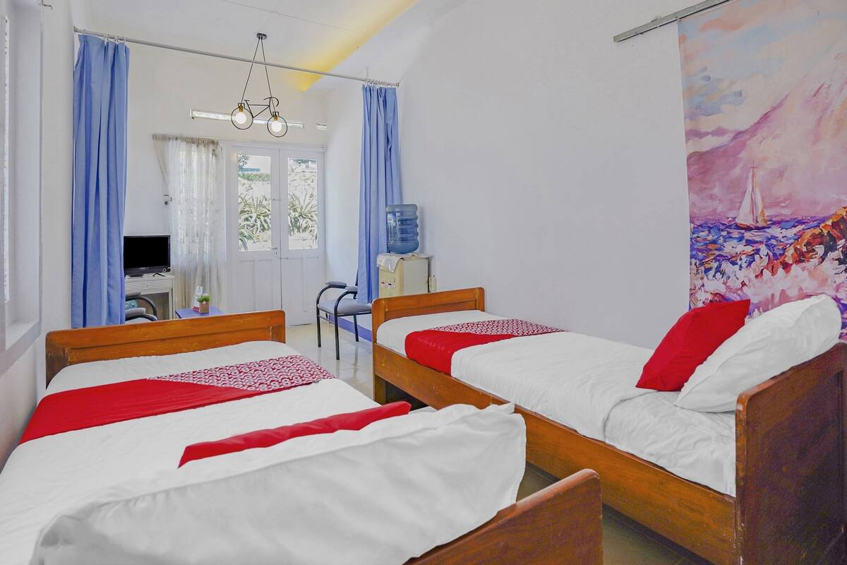 Quarto com duas camas de madeira, uma escrivaninha com cadeira, um dispensador de água e um quadro na parede.