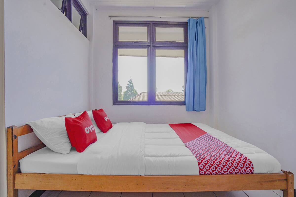 Uma cama de solteiro com estrutura de madeira, roupa de cama branca e almofadas vermelhas e brancas.