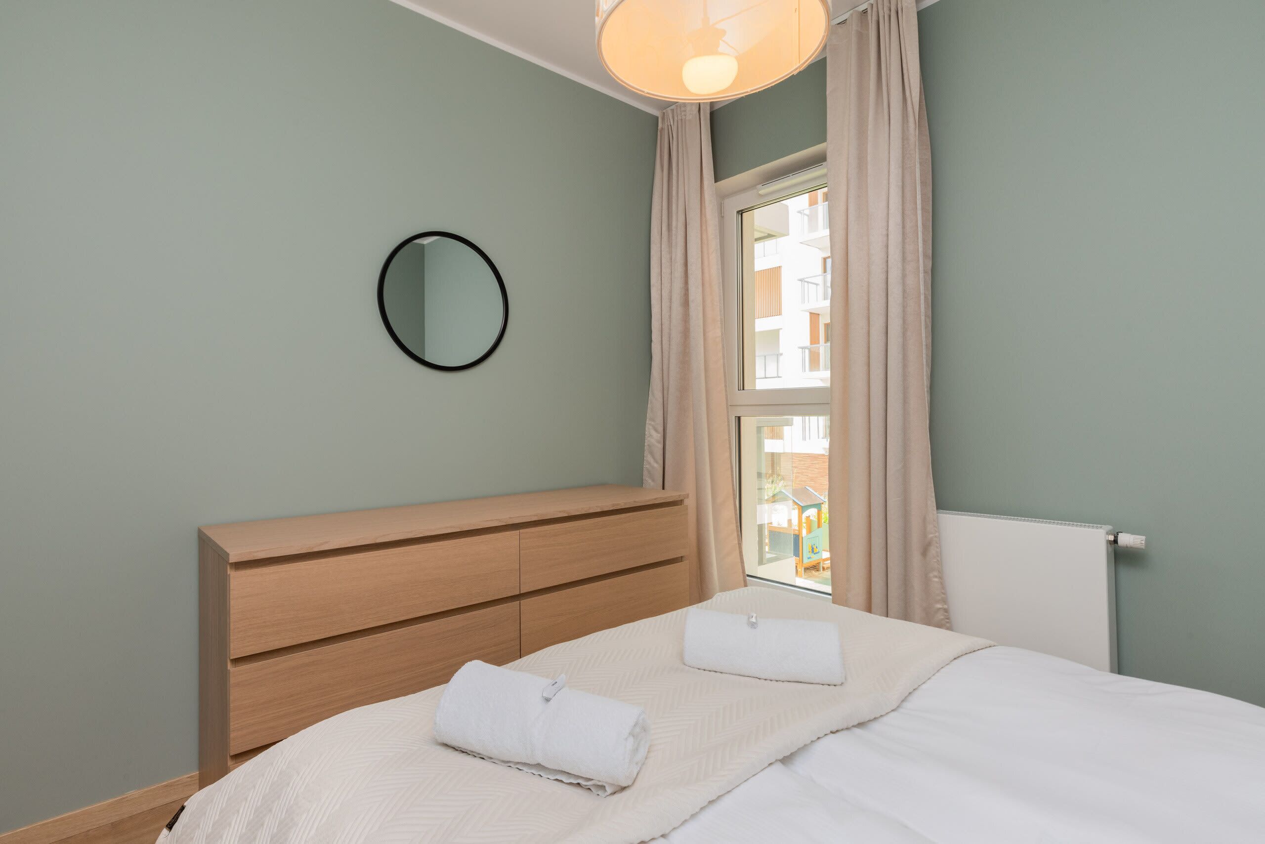 A bedroom with a bed, a bedside table, a mirror, and a window with curtains.