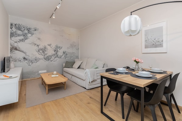 A modern living room with a dining area, featuring a sofa, a coffee table, a dining table with chairs, a TV, and artwork on the walls.