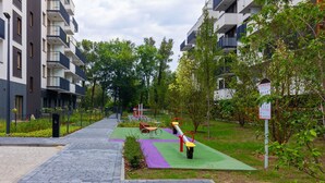 A pathway leading to a green outdoor area with exercise equipment, surrounded by trees and a multi-story building with balconies.