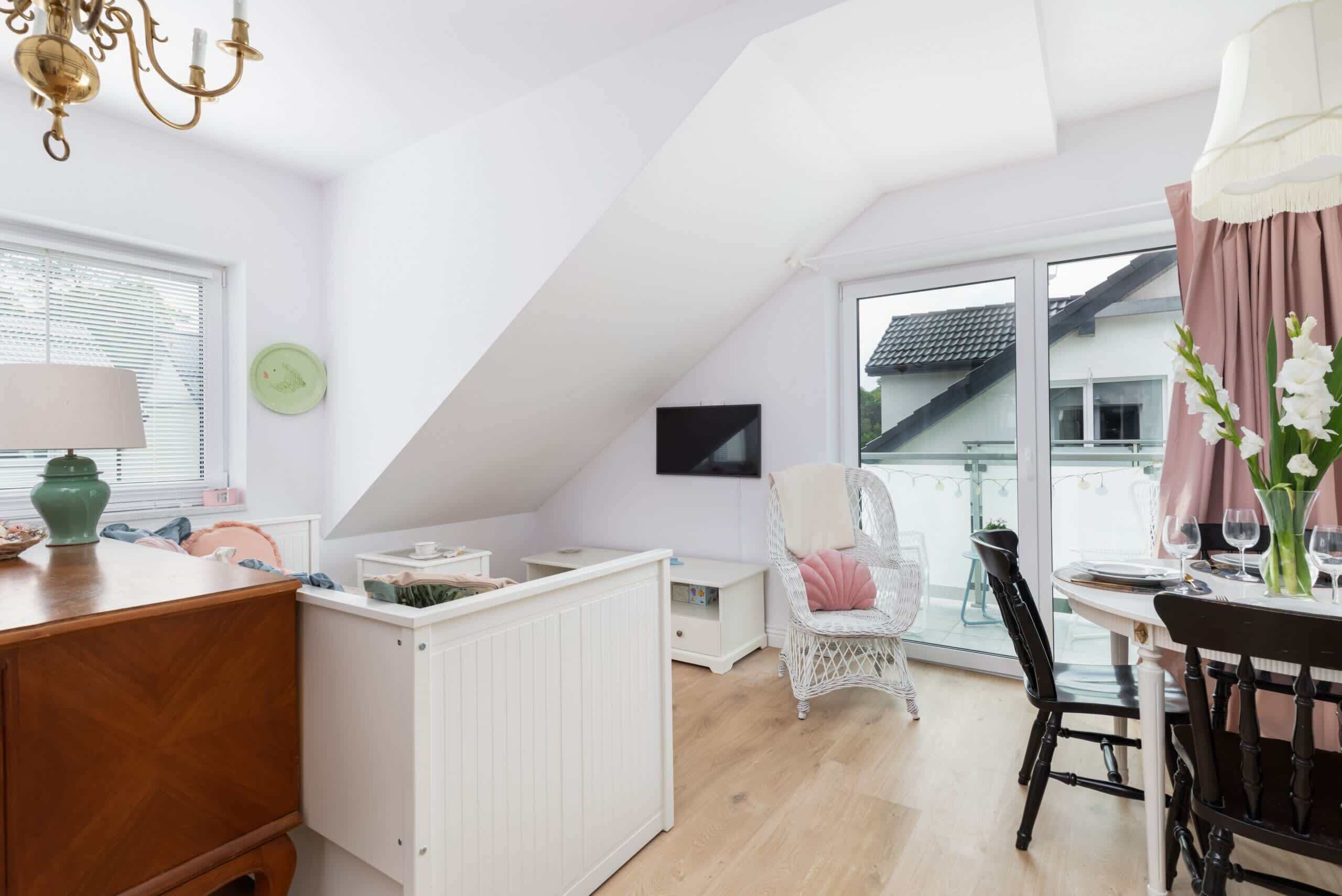 Une pièce chaleureuse avec une commode en bois, un radiateur blanc, un fauteuil en osier, une table à manger avec des chaises et une grande fenêtre avec des stores.