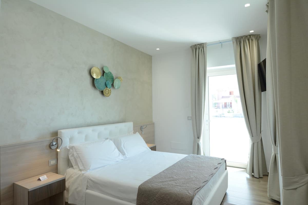 Ein Hotelzimmer mit einem großen Bett, einem Nachttisch, einer Wandleuchte und Blick durch Vorhänge ins Freie.