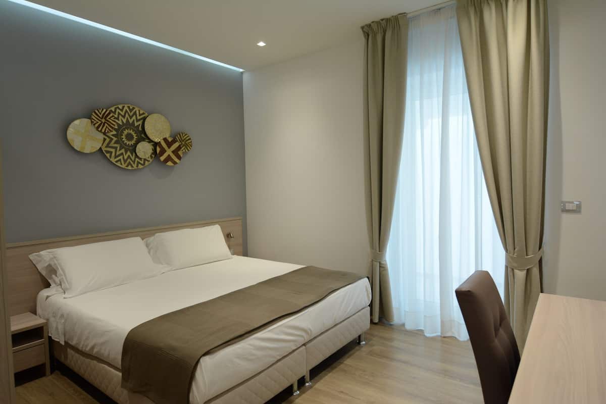 Ein modernes Hotelzimmer mit einem großen Bett, einem Holfnachtkästchen, einem Schreibtisch und einem Fenster mit Vorhängen.