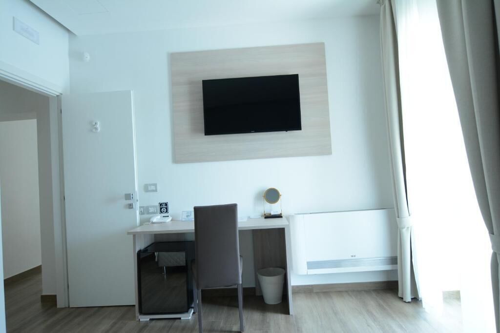 A modern hotel room with a flat-screen TV mounted on the wall, a desk with a chair, a small round mirror, and a radiator.