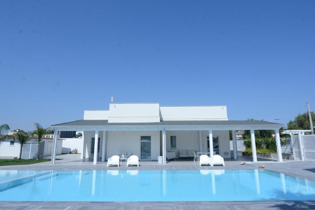 Una casa moderna con piscina, patio cubierto y mobiliario blanco.