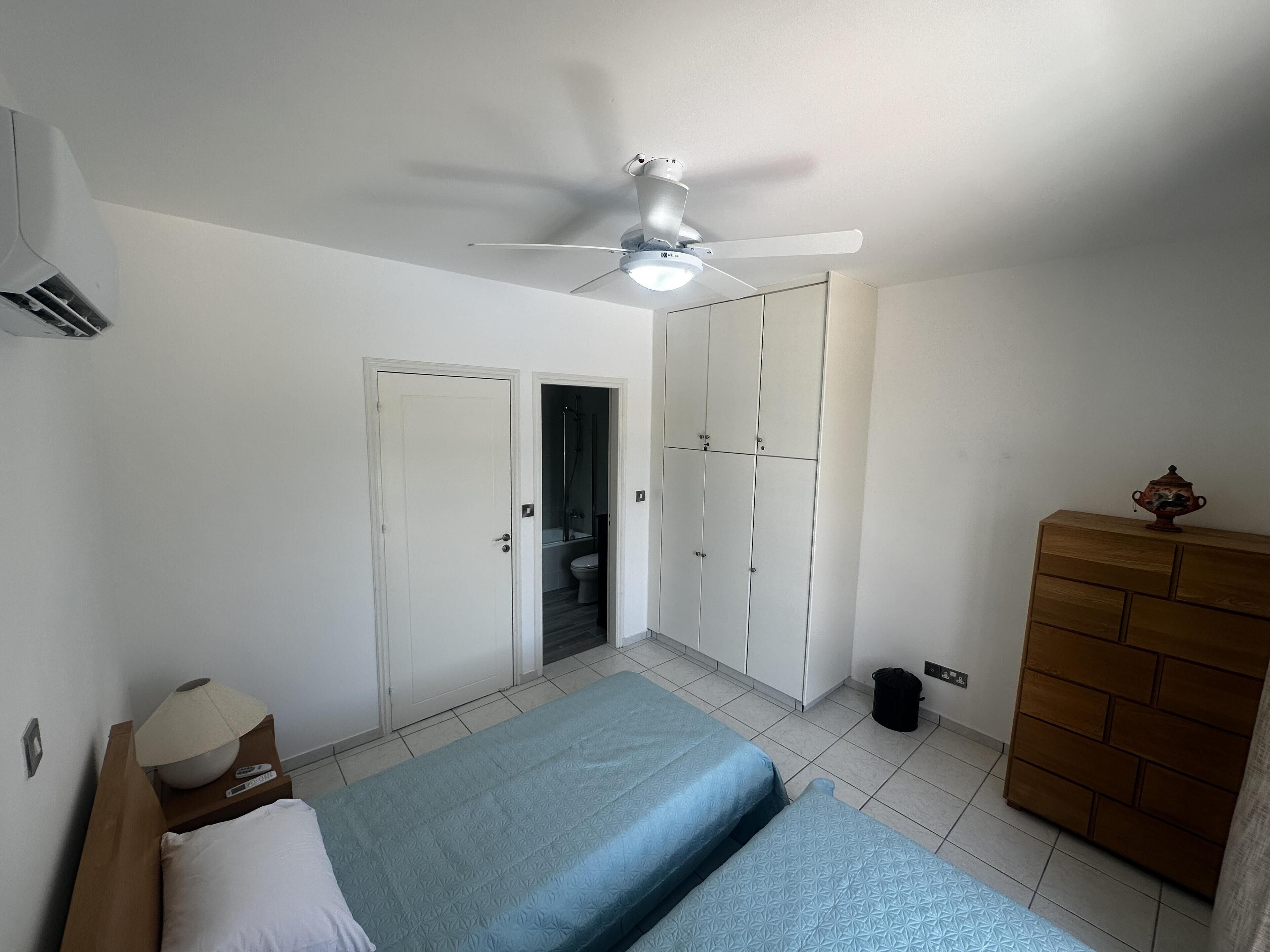 A bedroom with a bed, wardrobe, ceiling fan, and a small table with a lamp.