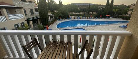 Een balkon met een houten tafel en stoelen, met uitzicht op een zwembad met ligbedden en parasols, omgeven door gebouwen en bomen.