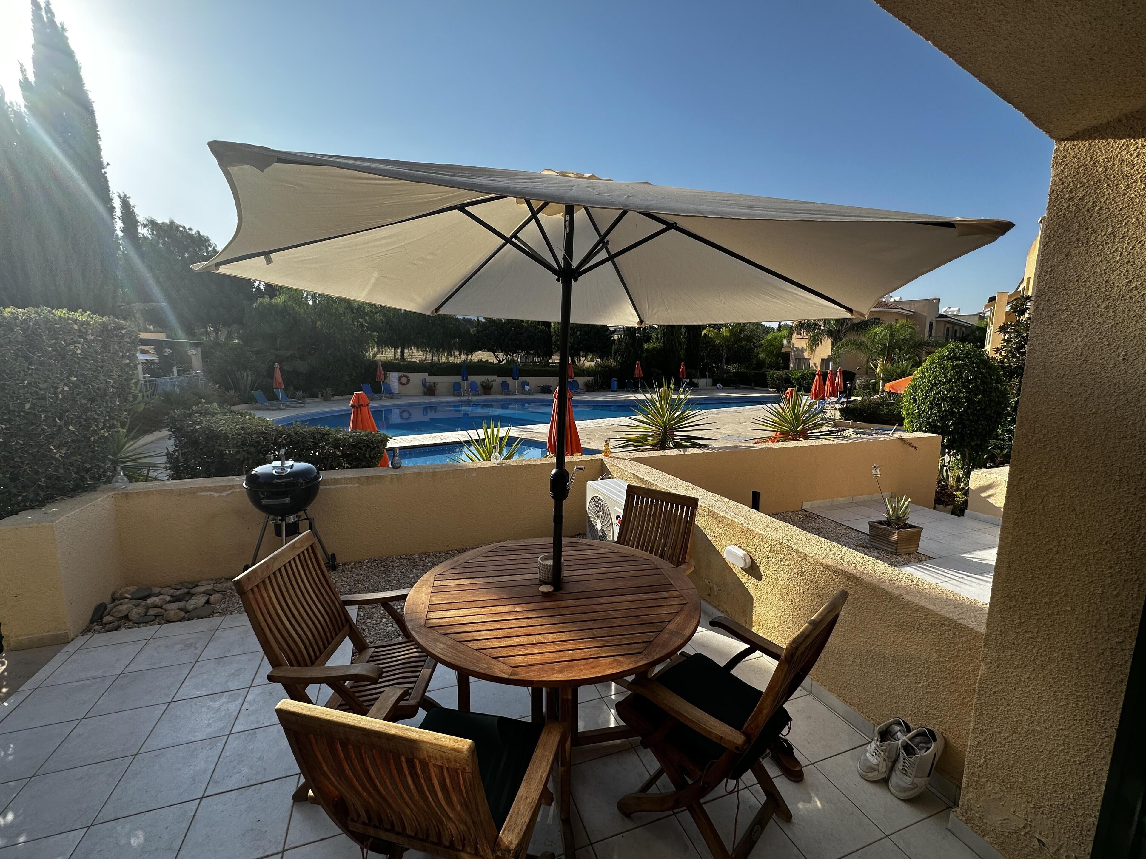 Une terrasse avec une table ronde en bois et des chaises, un parasol et une vue sur une piscine.