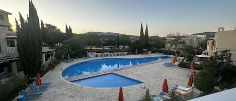 A swimming pool with lounge chairs and umbrellas, surrounded by trees and buildings.