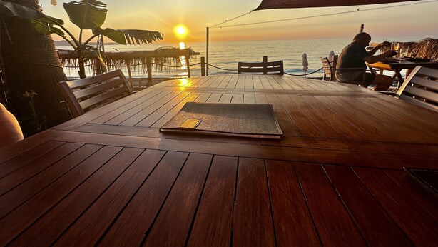 Une terrasse en bois avec une table et des bancs, offrant une vue sur l’océan au coucher du soleil.