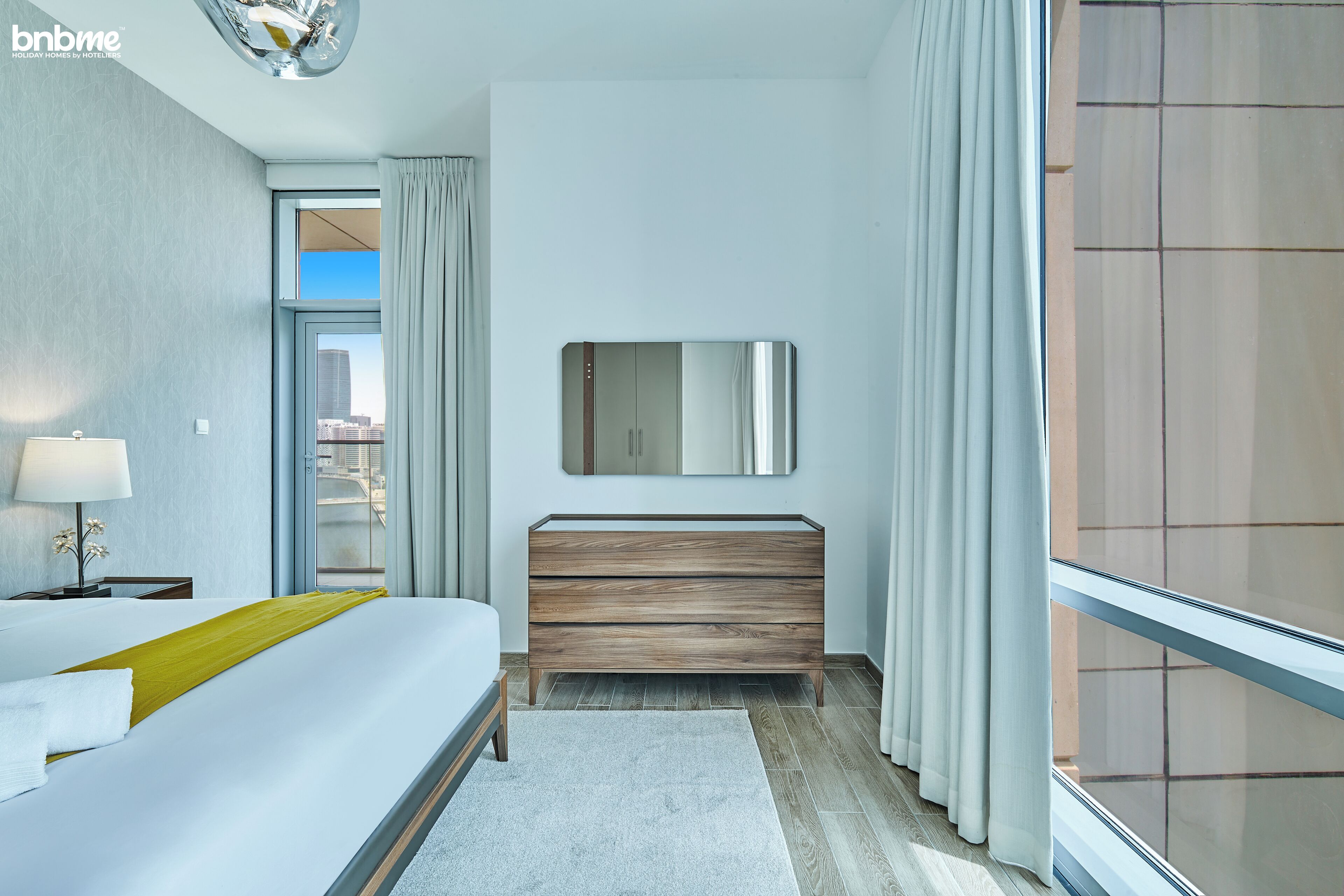 A modern hotel room with a bed, a wooden dresser, a mirror, and a view of the city through a large window.