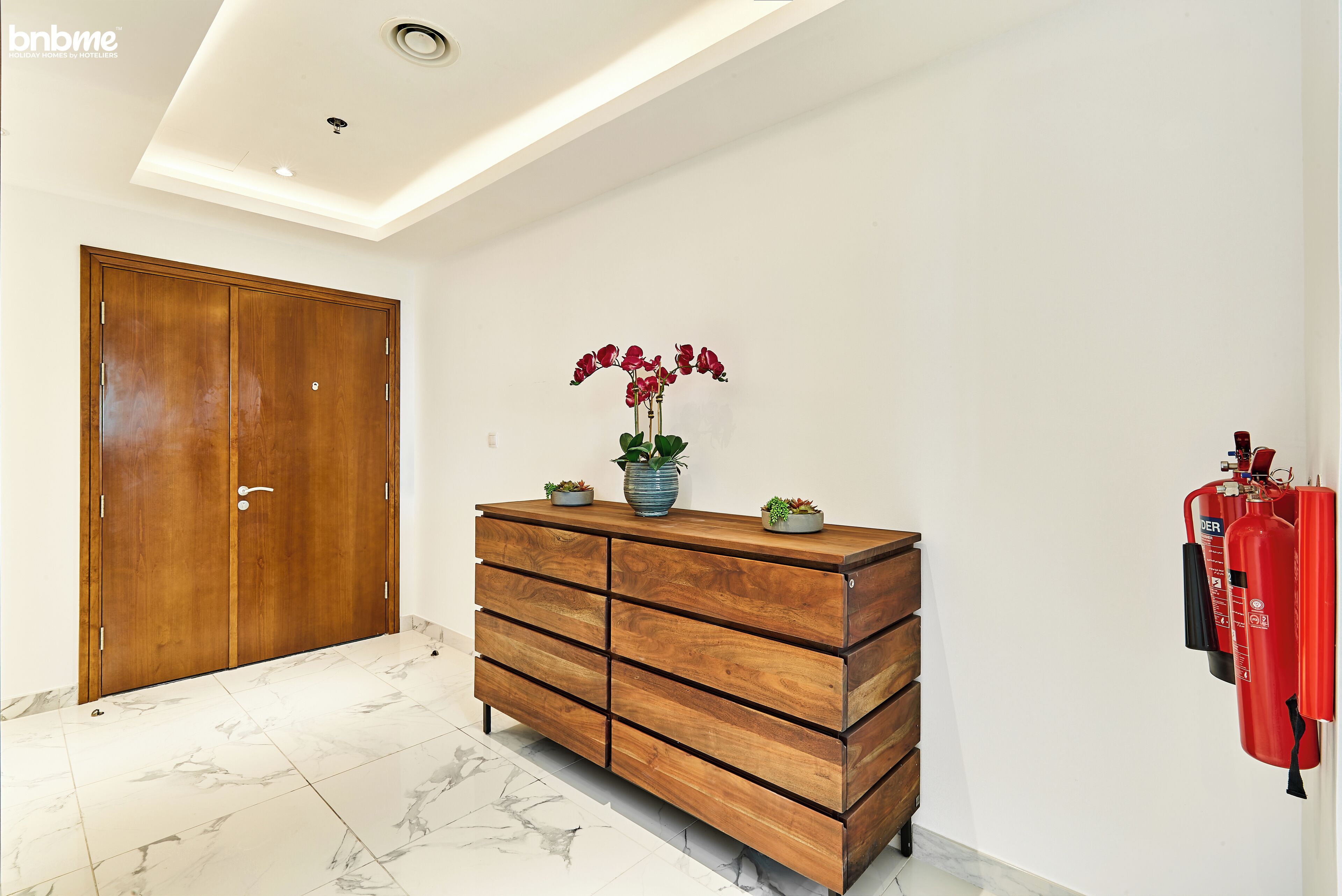 A room with a wooden wardrobe, a marble floor, a vase with flowers, and a fire extinguisher on the wall.