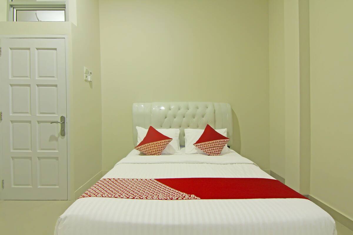 A hotel room with a white bed, red and white pillows, and a red throw blanket.