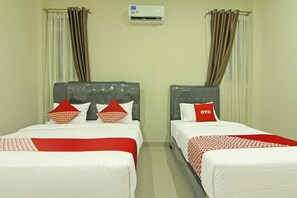 A hotel room with two beds, each with a grey headboard, white bedding, and red accents. An air conditioning unit is mounted on the wall above the beds.