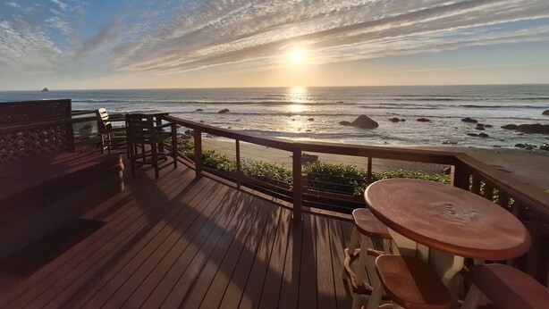 Um deck de madeira com mesa e bancos, com vista para o oceano ao pôr do sol.