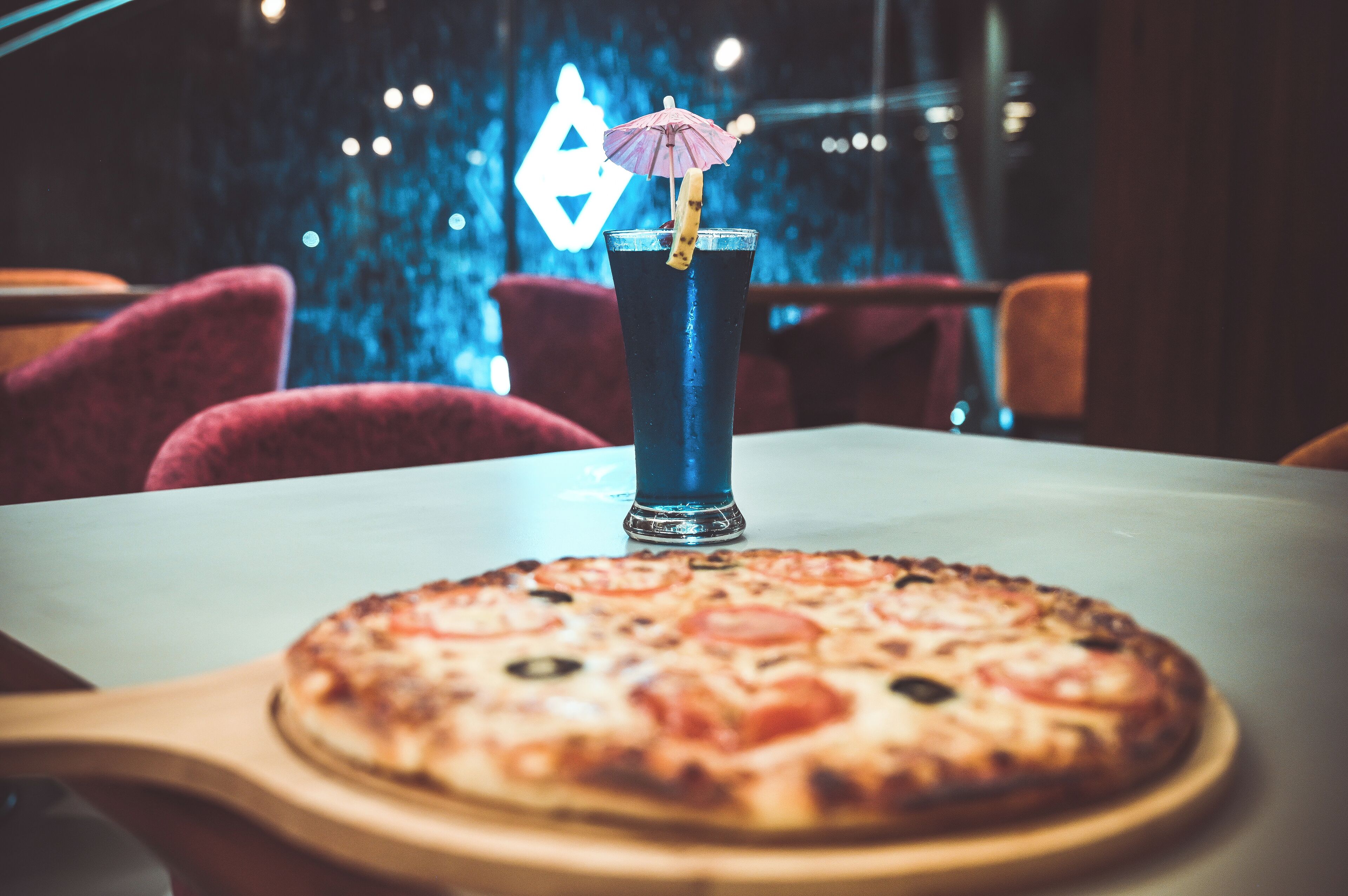 Una pizza sobre una tabla de madera con una bebida azul en un vaso, con pajita y un paraguas pequeño en la parte superior.