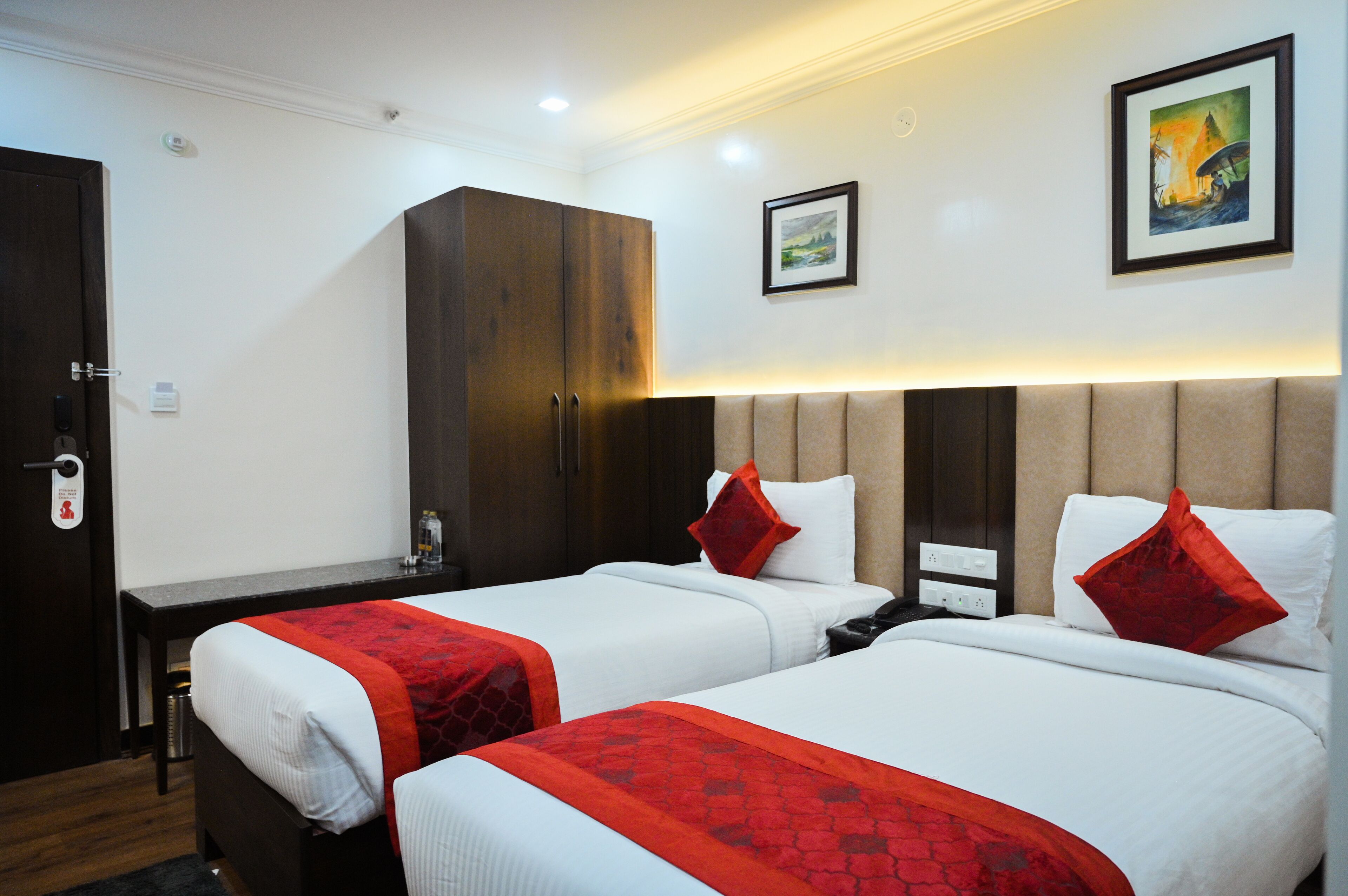 Quarto de hotel com duas camas, cada uma com colcha vermelha e branca, um guarda-roupa de madeira, uma escrivaninha com cadeira e um quadro emoldurado na parede.