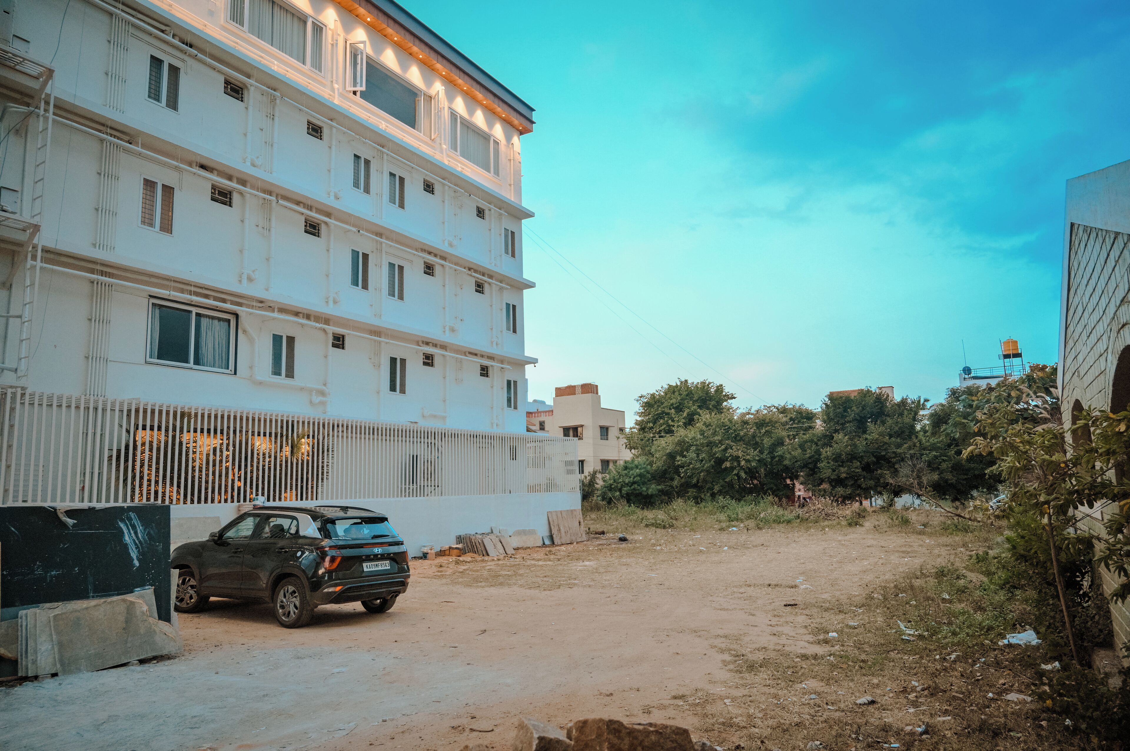 A multi-story building with a white facade, a parked SUV, and a dirt ground area with some vegetation.