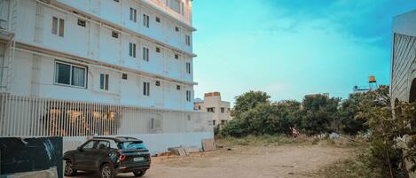 Un edificio a più piani con facciata bianca, un SUV parcheggiato e un’area a terra sterrata con della vegetazione.