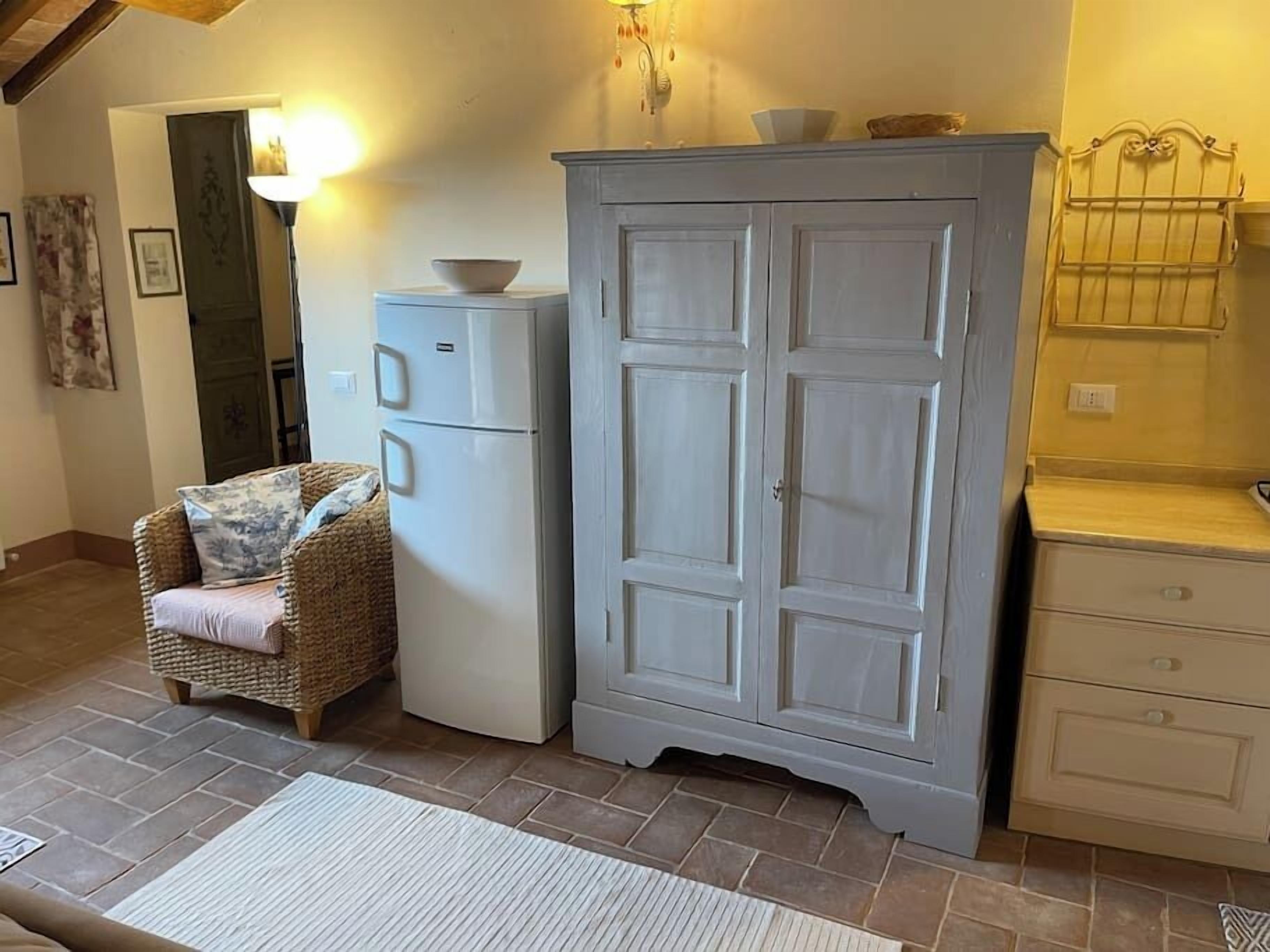 A room with a wicker chair, a white refrigerator, a large grey cabinet, and a small white dresser.