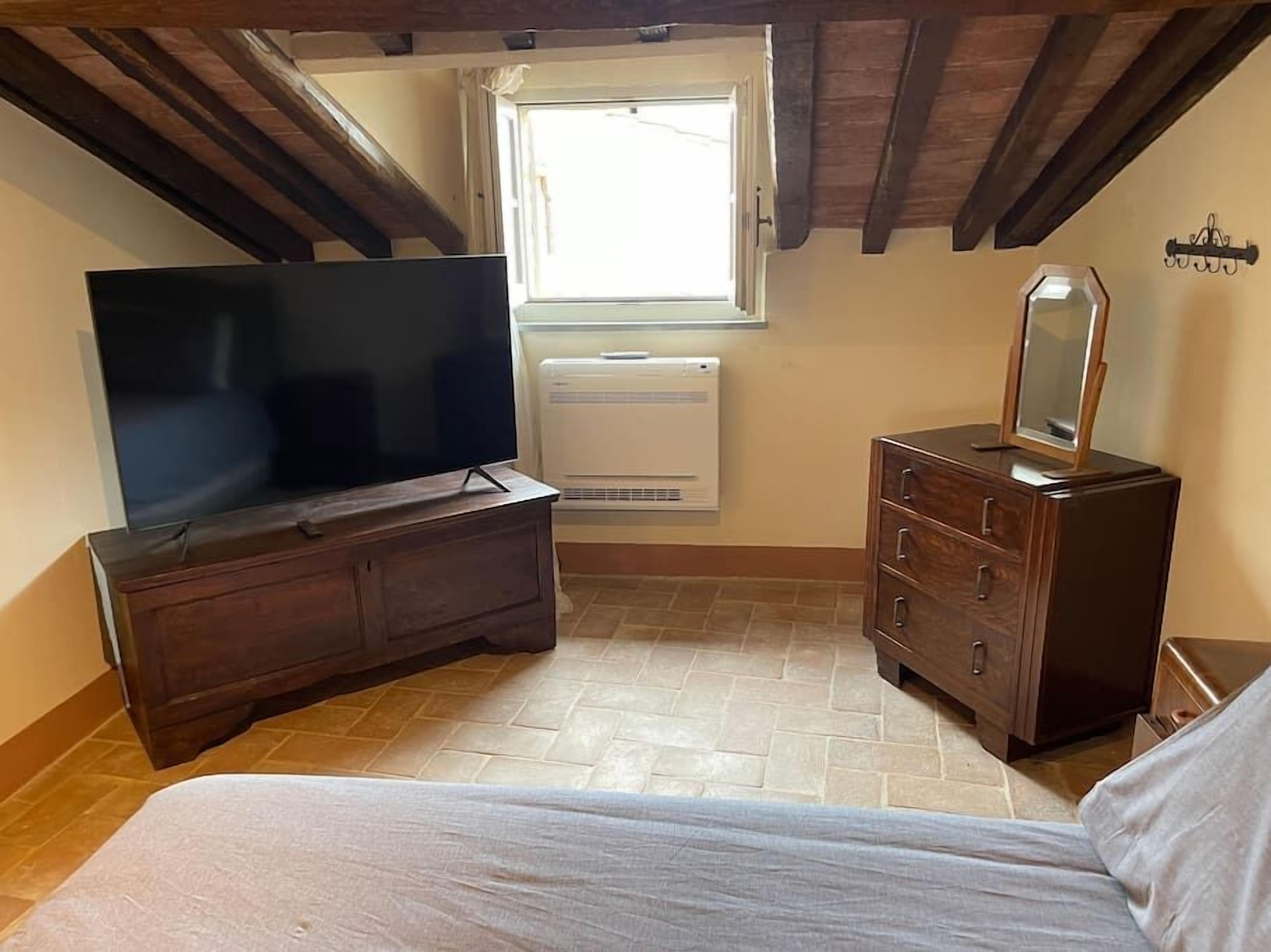 Une chambre à coucher avec un grand téléviseur à écran plat posé sur une commode en bois, une petite fenêtre donnant sur un paysage agréable, un miroir sur une autre commode et un lit recouvert d’une couverture blanche.