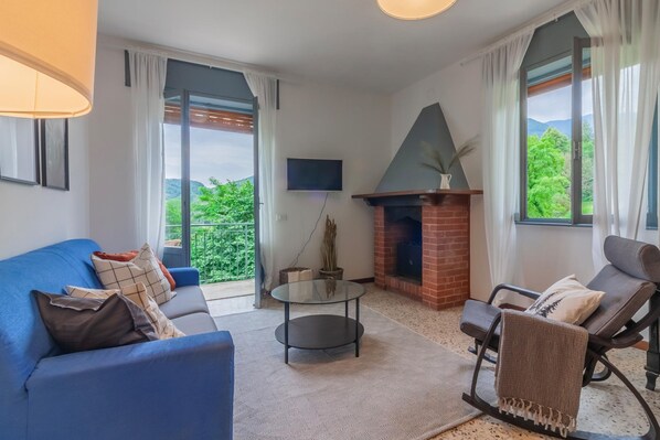 Un salon avec un canapé bleu, une table basse en verre, une cheminée et une vue sur la verdure grâce à de grandes fenêtres.
