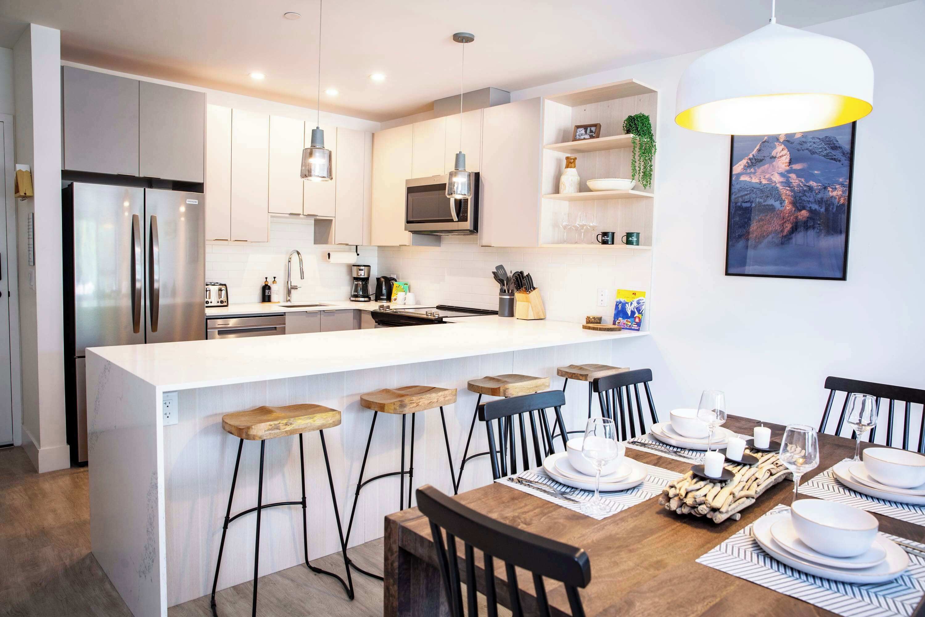 Una cocina moderna con una isla central, taburetes de bar, una mesa de comedor para cuatro personas y una lámpara colgante.