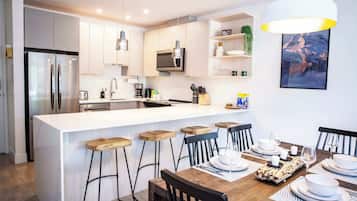 Una cocina moderna con una isla central, taburetes de bar, una mesa de comedor para cuatro personas y una lámpara colgante.
