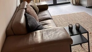Ein modernes Wohnzimmer mit einer Ledersofa, einem kleinen Tisch mit Gläsern und einer Küchenzeile mit Kühlschrank.