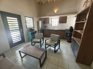 Un espace de vie compact comprenant un coin cuisine, deux fauteuils, une table basse et une porte équipée de stores horizontaux.