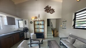 Ein modernes Wohnzimmer mit Sofa, Sessel und Couchtisch. Im Hintergrund ist eine Küchenzeile mit Schränken und Herd erkennbar.