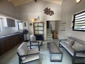 Ein modernes Wohnzimmer mit Sofa, Sessel und Couchtisch. Im Hintergrund ist eine Küchenzeile mit Schränken und Herd erkennbar.