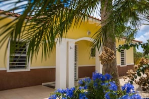 A yellow building with a number '24' on the door, palm trees, and blue flowers.