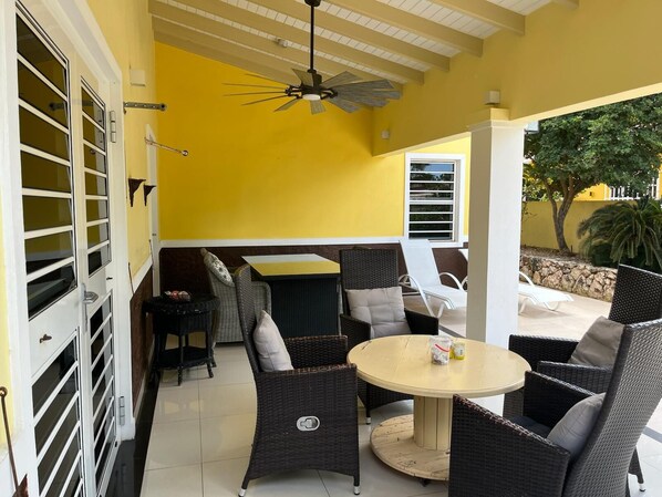 A room with a yellow wall, a ceiling fan, wicker furniture, and a window overlooking a garden.
