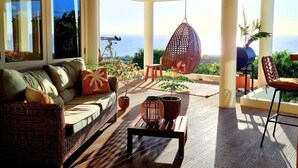 A sunlit patio with a wicker sofa, a hanging chair, and a table with potted plants. - Crown Terrace 18 by Bookaparadise (Kralendijk)